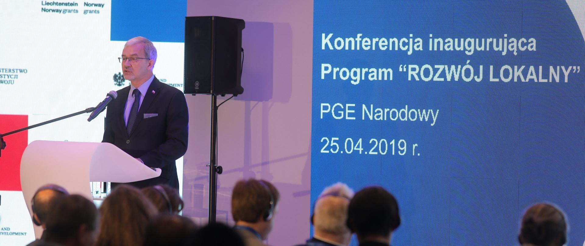 Na pierwszym planie widownia od tyłu, po lewej stronie minister Jerzy Kwieciński przy białej mównicy, na dalszym planie dwa ekrany w kolorach białym i niebieskim - jeden z logo ministerstwa oraz funduszy norweskich, drugi z nazwą wydarzenia: "Konferencja inaugurująca Program "Rozwój lokalny", PGE Narodowy, 25.04.2019 r.".