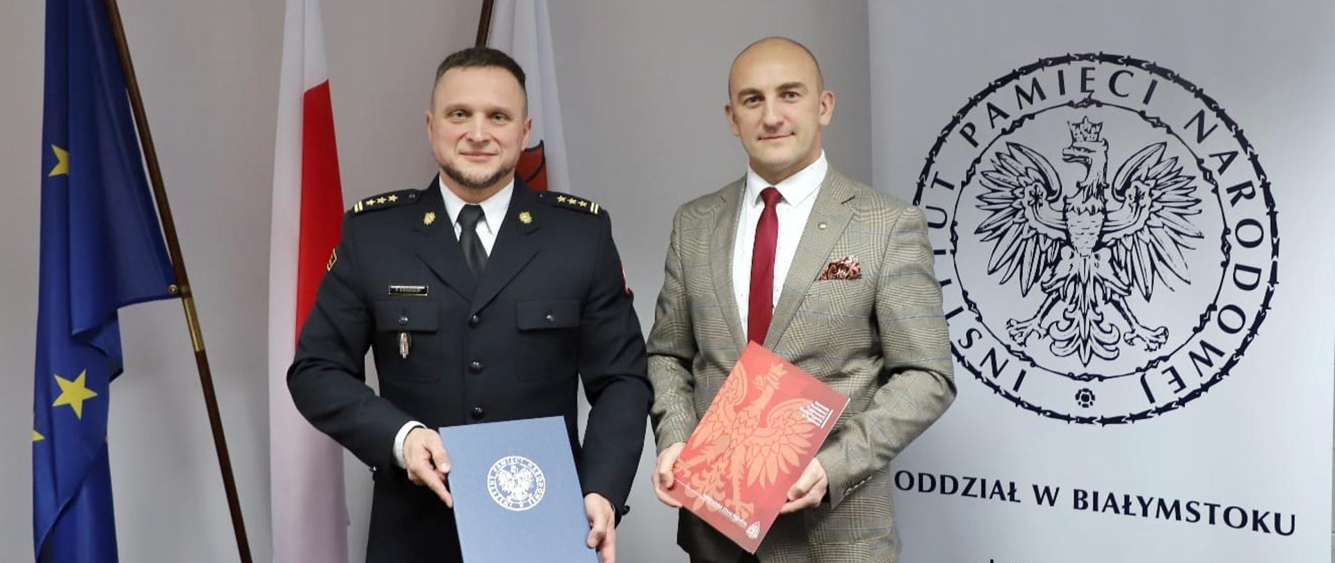 Dwie osoby stoją trzymają dokumenty w dłoni z tyłu za nimi flagi Polski i UE.
