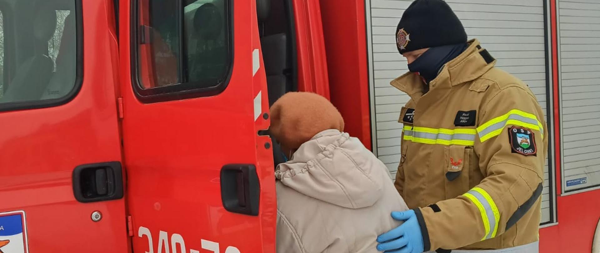 Strażak ochotniczej straży pożarnej w maseczce i rękawiczkach pomaga starszej osobie wsiąść do lekkiego pojazdu pożarniczego.
