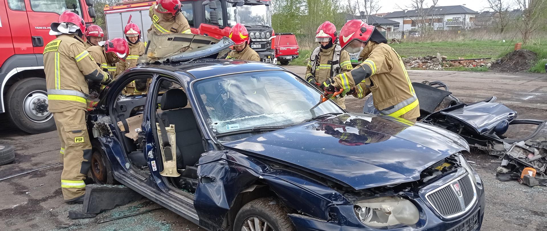 Na zdjęciu widać zniszczone auto osobowe koloru ciemnego. Strażacy- ratownicy w ubraniach specjalnych koloru piaskowego oraz czerwonych hełmach operują sprzętem hydraulicznym. Jeden strażak wycina przednią szybę auta. W tle widać dwa czerwone wozy strażackie.