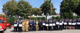 Strażacy PSP i OSP stoją razem do wspólnego zdjęcia, z lewej strony stoi czerwony wóz strażacki, w tle drzewa