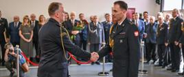 Na zdjęciu dwóch oficerów Państwowej Straży Pożarnej w galowych mundurach uściskiem dłoni dokonuje symbolicznego aktu przekazania obowiązków lub gratulacji. W tle zgromadzeni są goście i funkcjonariusze, niektórzy trzymają kwiaty. Uroczystość odbywa się w hali, przestrzeń oddzielona jest czerwonymi barierkami. Fotograf uwiecznia moment z prawej strony kadru.