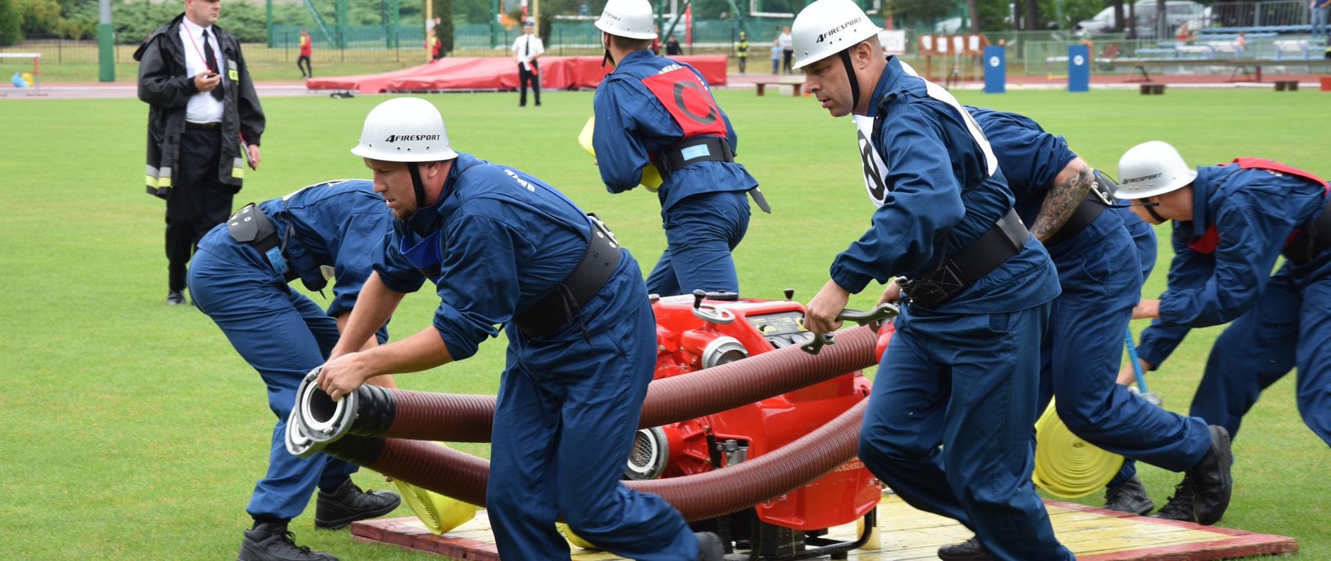 Na boisku strażacy biegną z odcinkami. Na drewnianym podeście stoi motopompa. W tle strażak trzyma w ręku stoper.