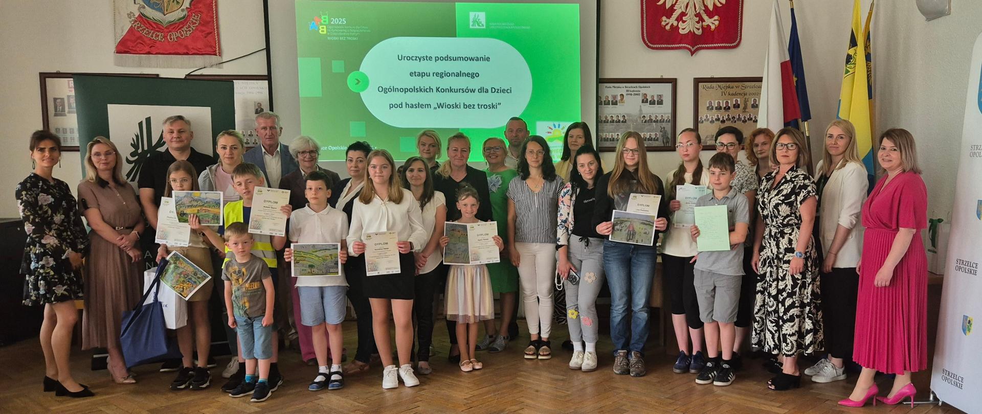 W reprezentacyjnej sali na tle ekranu z wyświetloną nazwą finału konkursów dla dzieci na tle flag, proporca z orłem białym, sztandaru Strzelec Opolskich i rollupu KRUS pozuje do zdjęcia duża grupa kobiet i mężczyzn. Przed nimi stoi dziewięcioro dzieci z dyplomami ze zdjęciami swoich prac w dłoniach. Wszyscy uśmiechają się do zdjęcia.