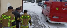 Dwóch strażaków w mundurach Ochotniczej Straży Pożarnej prowadzi starszego mężczyznę po szczepieniu do samochodu strażackiego.