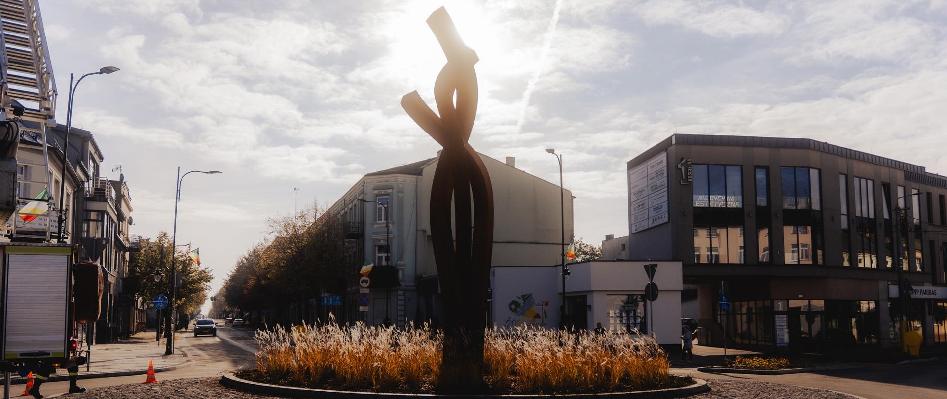 Zdjęcie, na pierwszym planie rzeźba "Splot", która przedstawia trzy przenikające się ramiona ze stali, w tle kamienice.