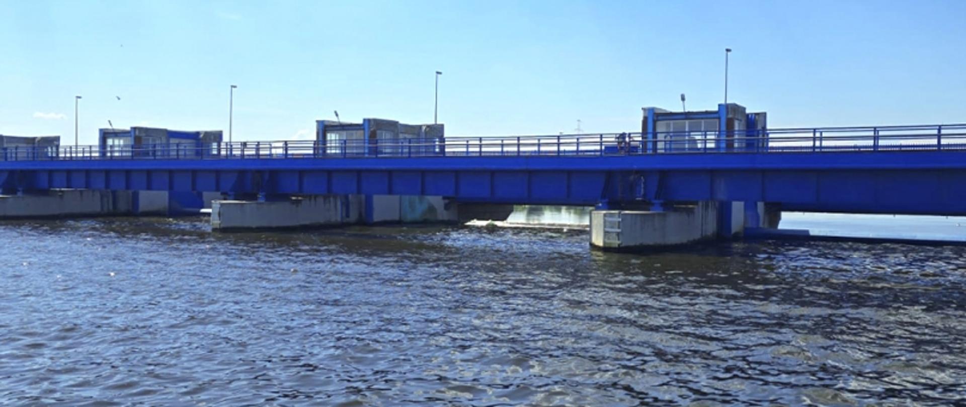 Most w kolorze niebieskim z barierkami i latarniami, rozciągający się nad wodą; na moście widoczne małe budynki techniczne.