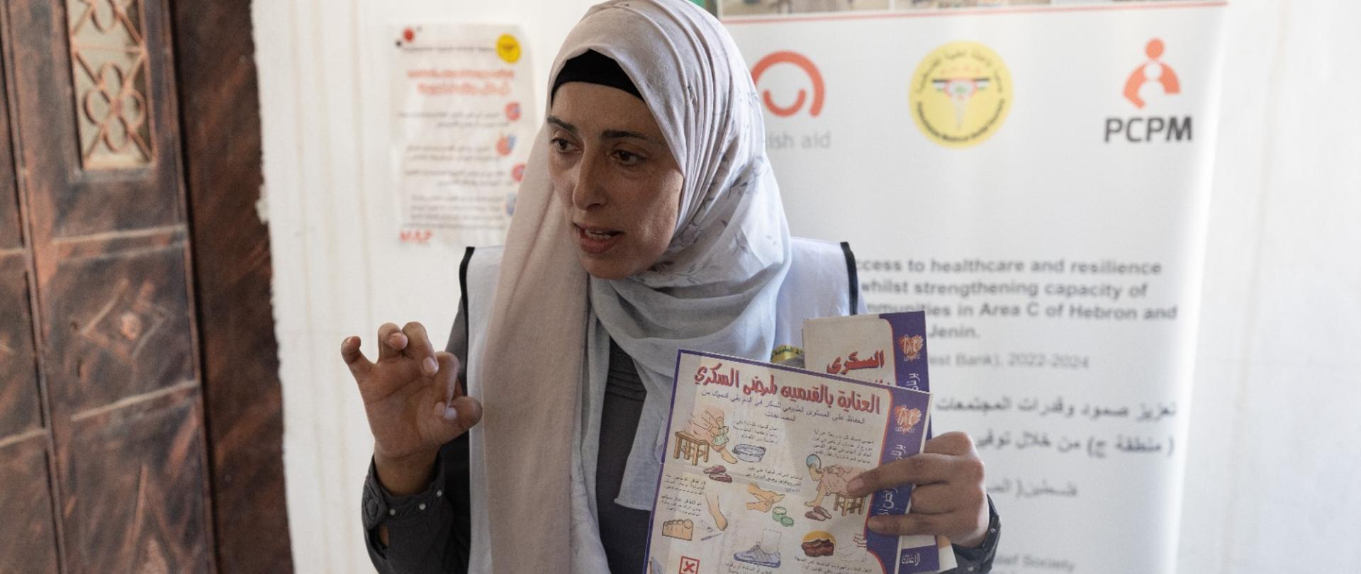 Kobieta z ulotką dotyczącą cukrzycy w alfabecie arabskim z rysunkami, w tle banner z logo Polska pomoc