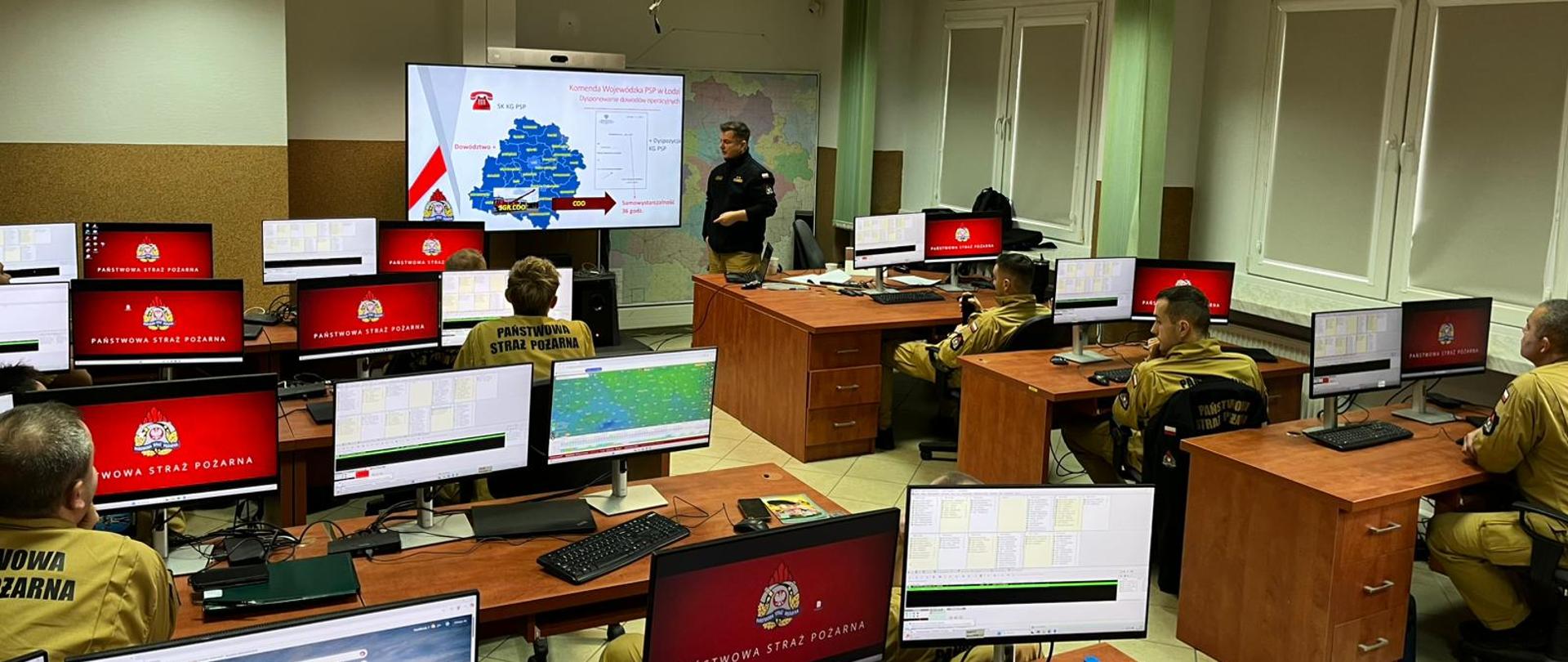 Strażacy w sali operacyjnej lub szkoleniowej z licznymi monitorami. Na centralnym ekranie wyświetlana jest mapa Polski z danymi. Jeden z dowódców stoi i prezentuje informacje.