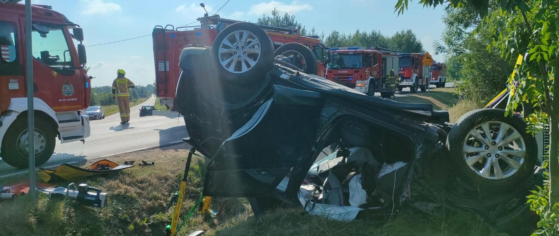 Zdjęcie przedstawia skutki wypadku samochodowego w słoneczny dzień. Na pierwszym planie ciemny SUV leży na dachu w rowie. Jego koła są skierowane do góry, a widoczne uszkodzenia sugerują poważne uderzenie. Po lewej stronie przewróconego pojazdu, na utwardzonej drodze, częściowo widoczny jest czerwony wóz strażacki. Dalej na drodze, w szeregu, stoi kilka innych pojazdów ratunkowych, w tym kolejne wozy strażackie. Strażak w żółto-ciemnym mundurze stoi przy pierwszym wozie strażackim, patrząc w kierunku miejsca zdarzenia. Po lewej stronie widoczny jest zielony znak drogowy z białym górnym paskiem i białym tekstem, prawdopodobnie wskazujący nazwę miejscowości „Zdonków” . Niebo jest czyste, z kilkoma białymi chmurami, a wzdłuż drogi i w tle rosną drzewa i rośliny. W pobliżu przewróconego samochodu widać porozrzucane szczątki.