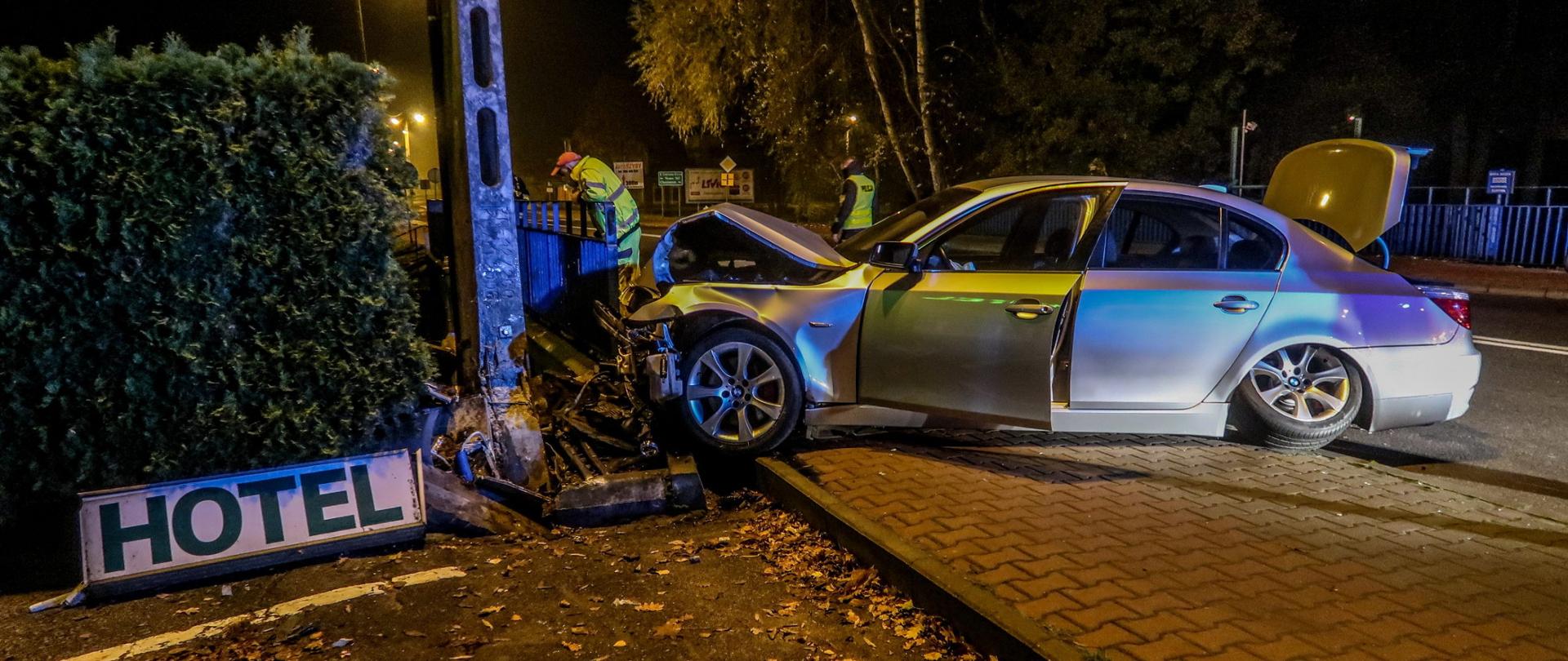 Wrak samochodu po uderzeniu w słup. W tle, pracownik pomocy drogowej oraz policjant - oceniają zniszczenia.
