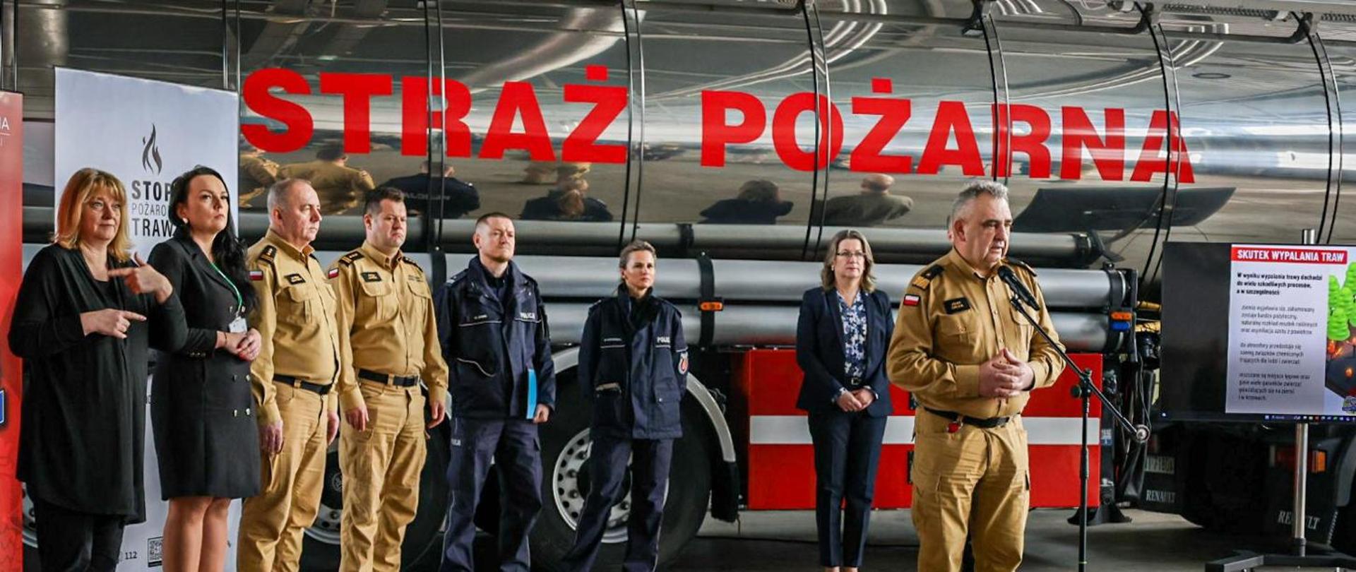Zastępca komendanta głównego PSP ubrany w strój strażacki koloru piaskowego, przemawia do mikrofonu. Za przemawiającym zastępcą komendanta na tle strażackiej cysterny z napisem Straż Pożarne stoją pozostali uczestnicy wydarzenia. Od lewej strony dwie kobiety w strojach cywilnych koloru czarnego dwaj przedstawiciele straży pożarnej w strojach strażackich koloru piaskowego, dwaj przedstawiciela policji w strojach policyjnych koloru granatowego mężczyzna i kobieta oraz od prawej strony stoi kobieta w ubraniu cywilnym koloru czarnego.