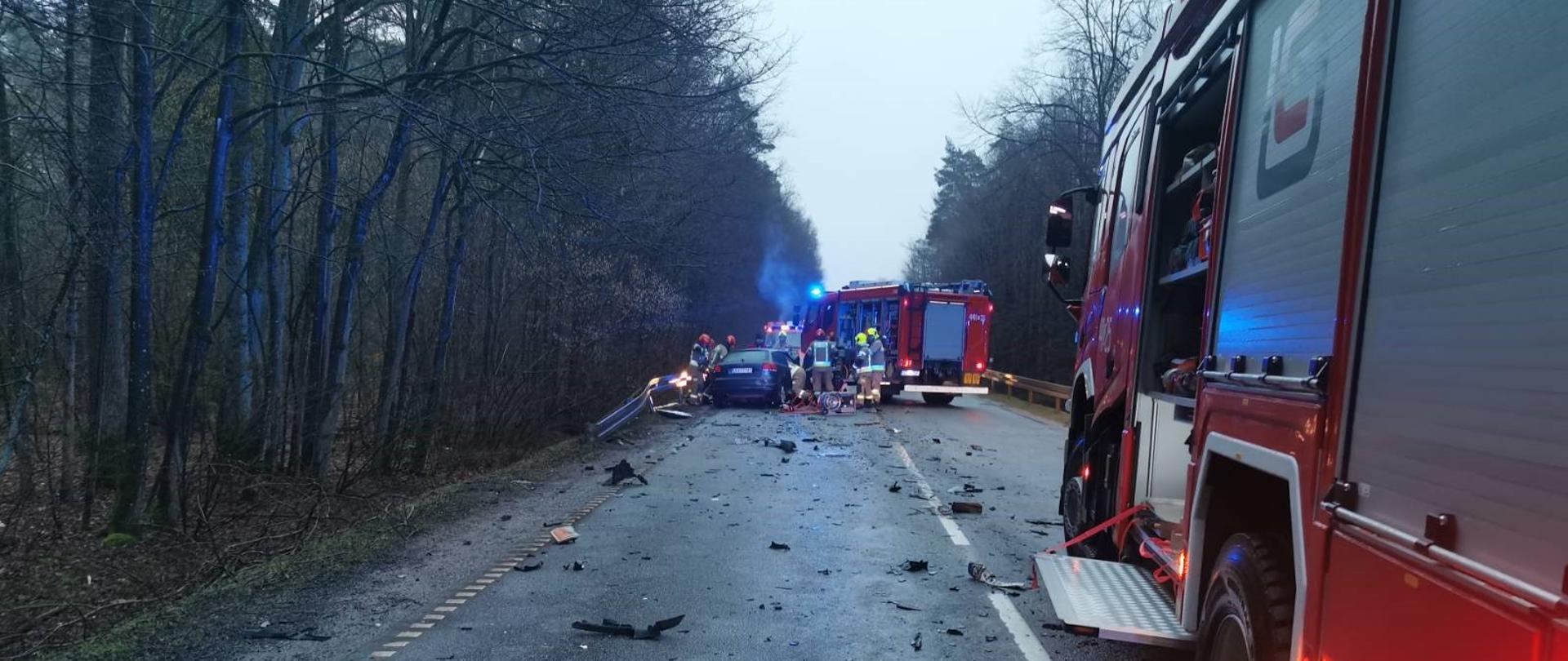 Samochód osobowy, który uległ wypadkowi, wokół znajdują się strażacy i wozy strażackie.