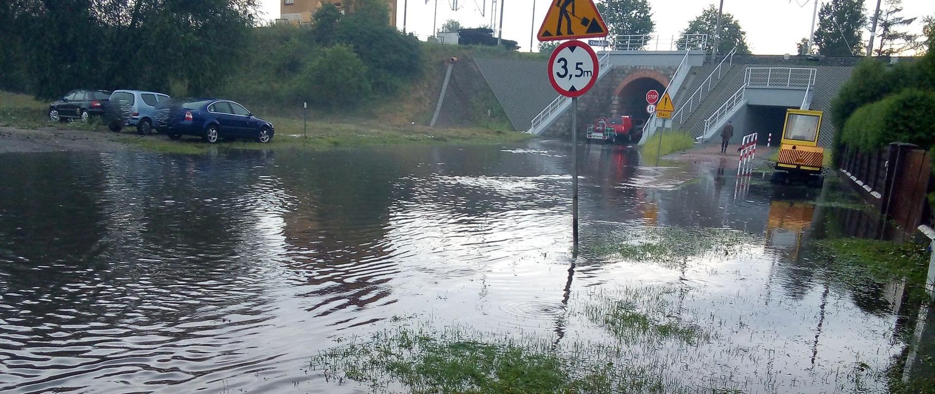 Na zdjęciu widać centrum miasta Chodzież po przejściu nawałnicy w 2020 roku. W wyniku bardzo intensywnych opadów deszczu zalana jest ulica (jezdnia), chodniki, posesje. W tyle wiadukt kolejowy, budynek PKP, samochody osobowe oraz samochód specjalny Państwowej Straży Pożarnej z motopompą pożarniczą dużej wydajności. 