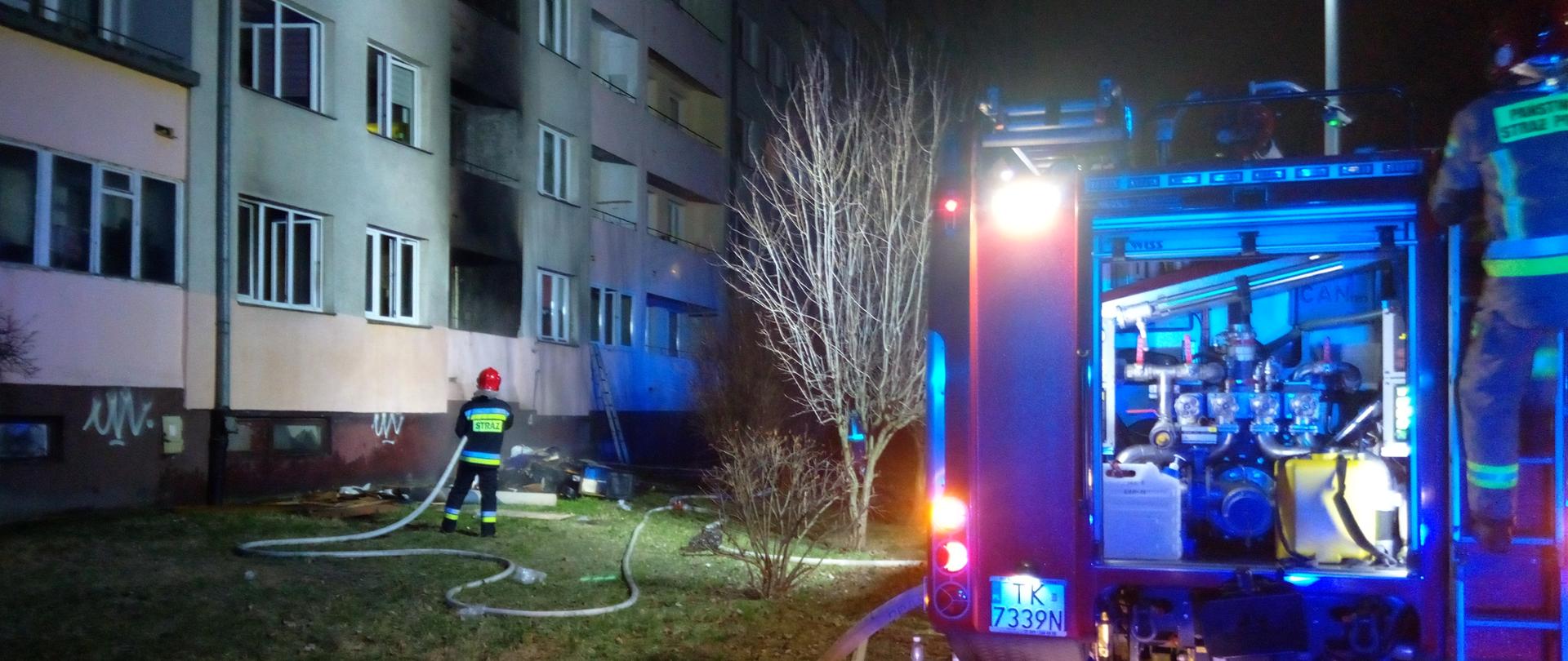 Zdjęcie przedstawia samochód pożarniczy stojący przed blokiem w którym doszło do pożaru. Widać strażaka który stoi z linią gaśniczą przed okopconymi balkonami. Nie widać ognia i dymu.