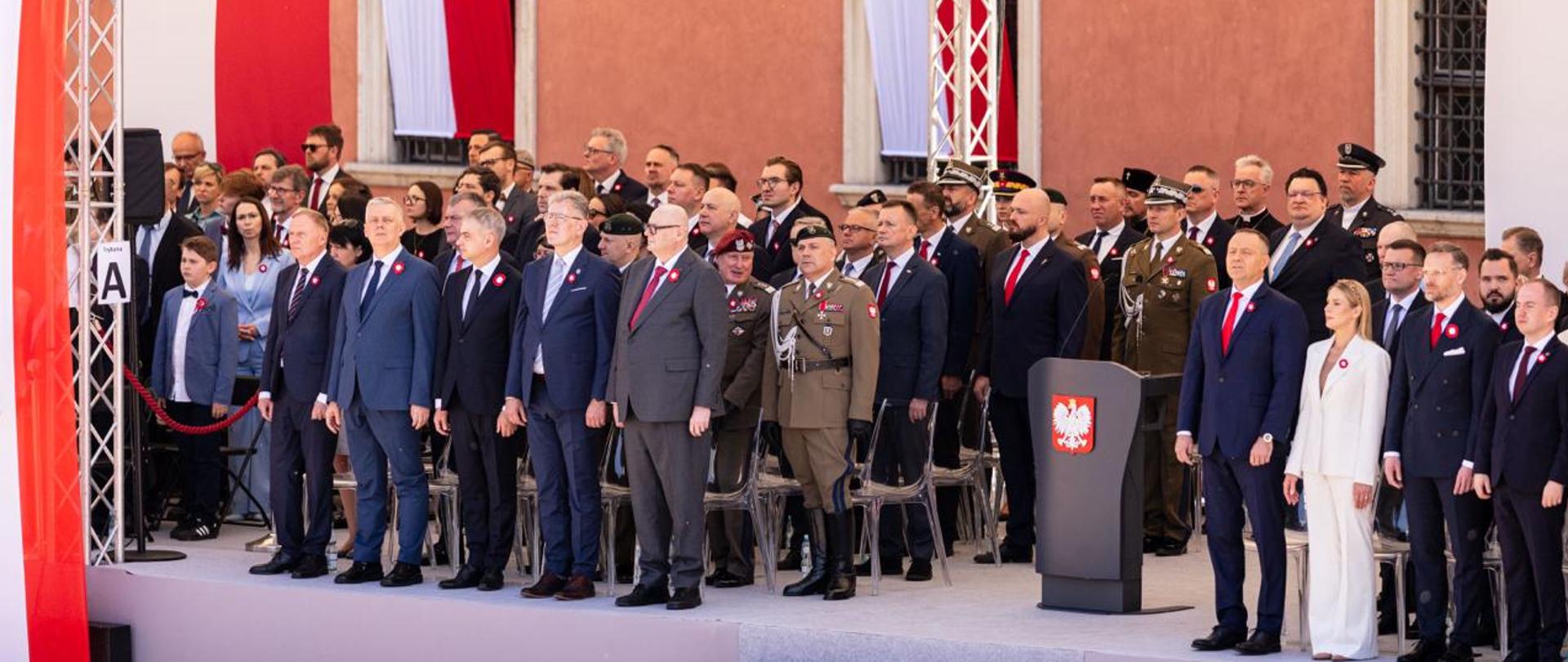 Na zdjęciu uroczystości państwowe z okazji 235. rocznicy uchwalenia Konstytucji 3 Maja. Na zdjęciu trybuna honorowa z najważniejszymi gośćmi. Po prawej stronie zdjęcia w pierwszym rzędzie widać parę prezydencką Karola i Martę Nawrockich. Na środku szara mównica z godłem (orzeł). Po lewej stronie od mównicy m.in. minister Tomasz Siemoniak, minister Krzysztof Gawkowski, wiceminister Czesław Mroczek. W tle zdjęcia front Zamku Królewskiego w Warszawie (widać trzy okna).Widać również wiszące biało-czerwone barwy narodowe. 