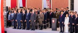 Na zdjęciu uroczystości państwowe z okazji 235. rocznicy uchwalenia Konstytucji 3 Maja. Na zdjęciu trybuna honorowa z najważniejszymi gośćmi. Po prawej stronie zdjęcia w pierwszym rzędzie widać parę prezydencką Karola i Martę Nawrockich. Na środku szara mównica z godłem (orzeł). Po lewej stronie od mównicy m.in. minister Tomasz Siemoniak, minister Krzysztof Gawkowski, wiceminister Czesław Mroczek. W tle zdjęcia front Zamku Królewskiego w Warszawie (widać trzy okna).Widać również wiszące biało-czerwone barwy narodowe. 