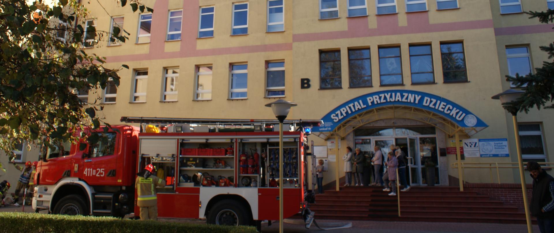 Zdjęcie przedstawia ewakuowany personel przed budynkiem szpitala, w tle wóz ratowniczo-gaśniczy 
