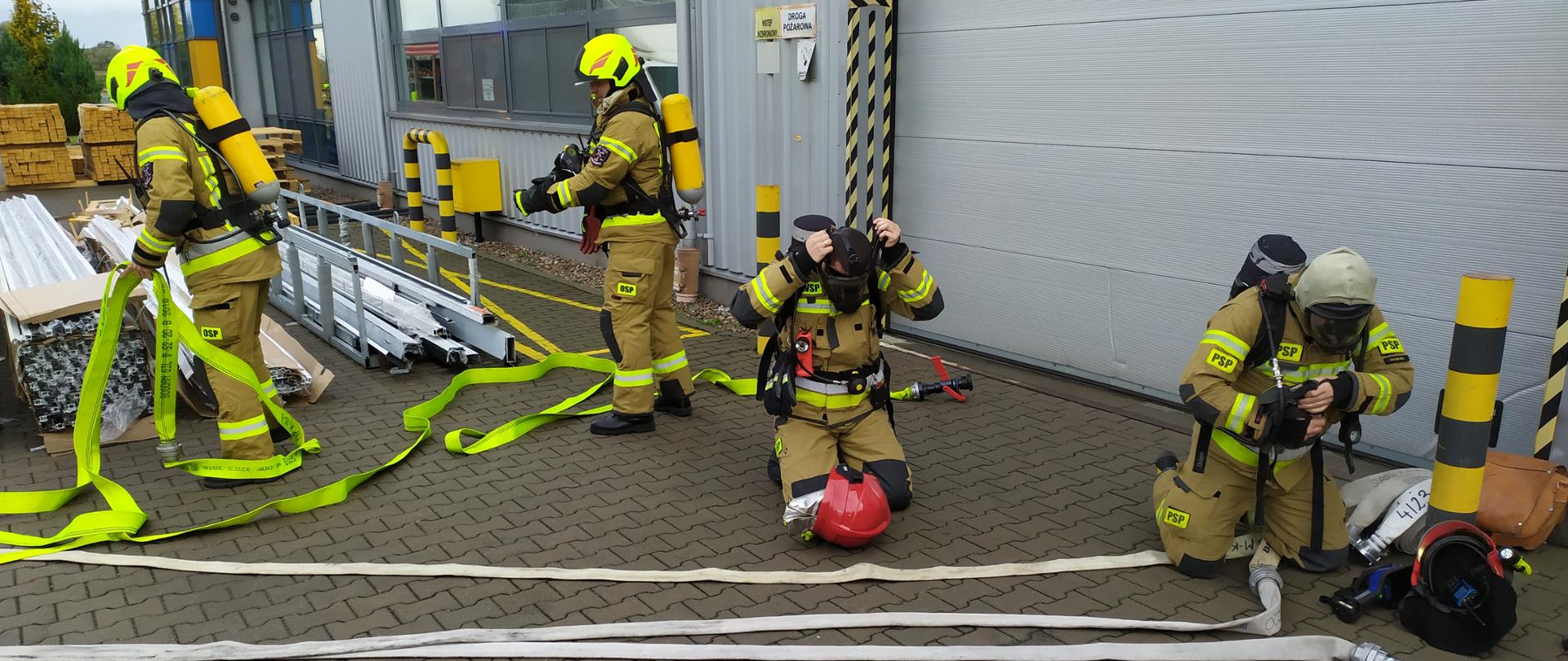 Strażacy rozwijają linie wężowe oraz podpinają sprzęt ochrony dróg oddechowych przy hali produkcyjnej