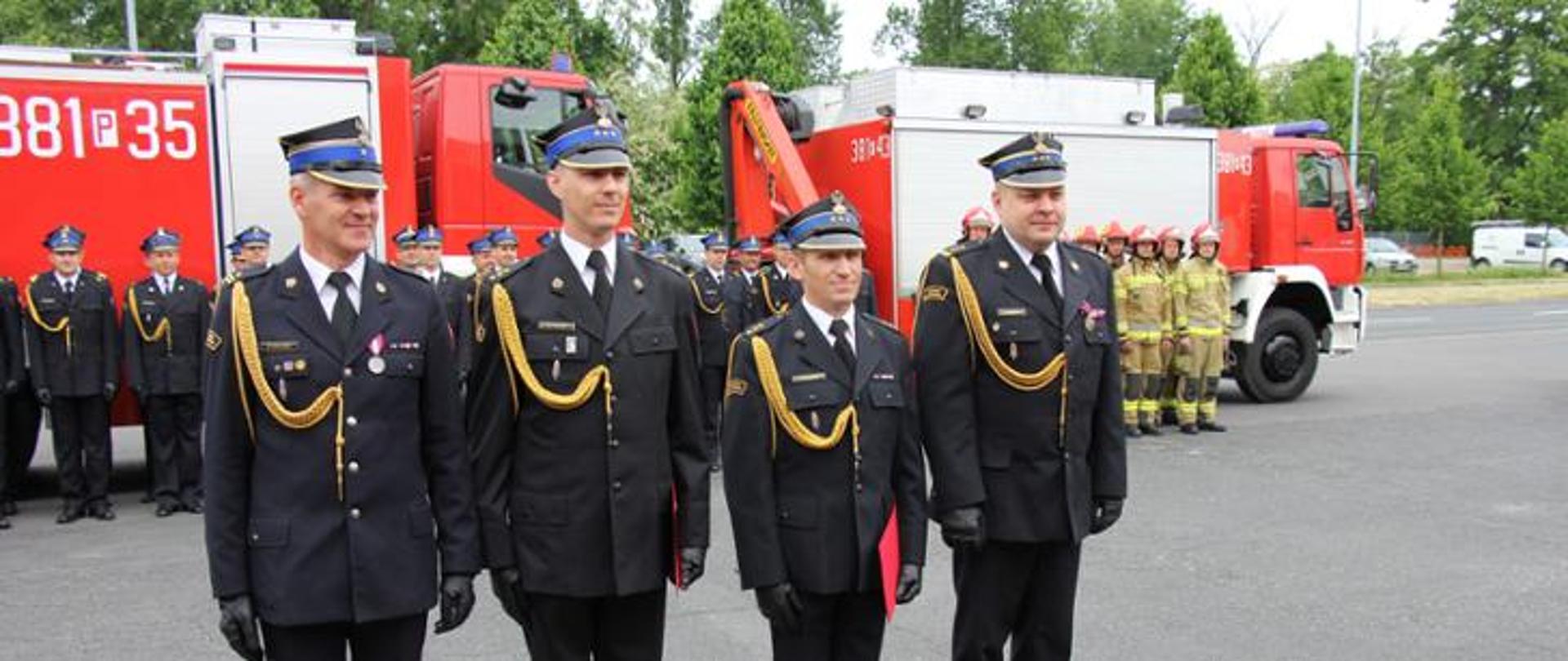 czterech strażaków w rogatywkach i mundurach ze sznurem stoi na baczność w szeregu, w tle pojazdy pożarnicze