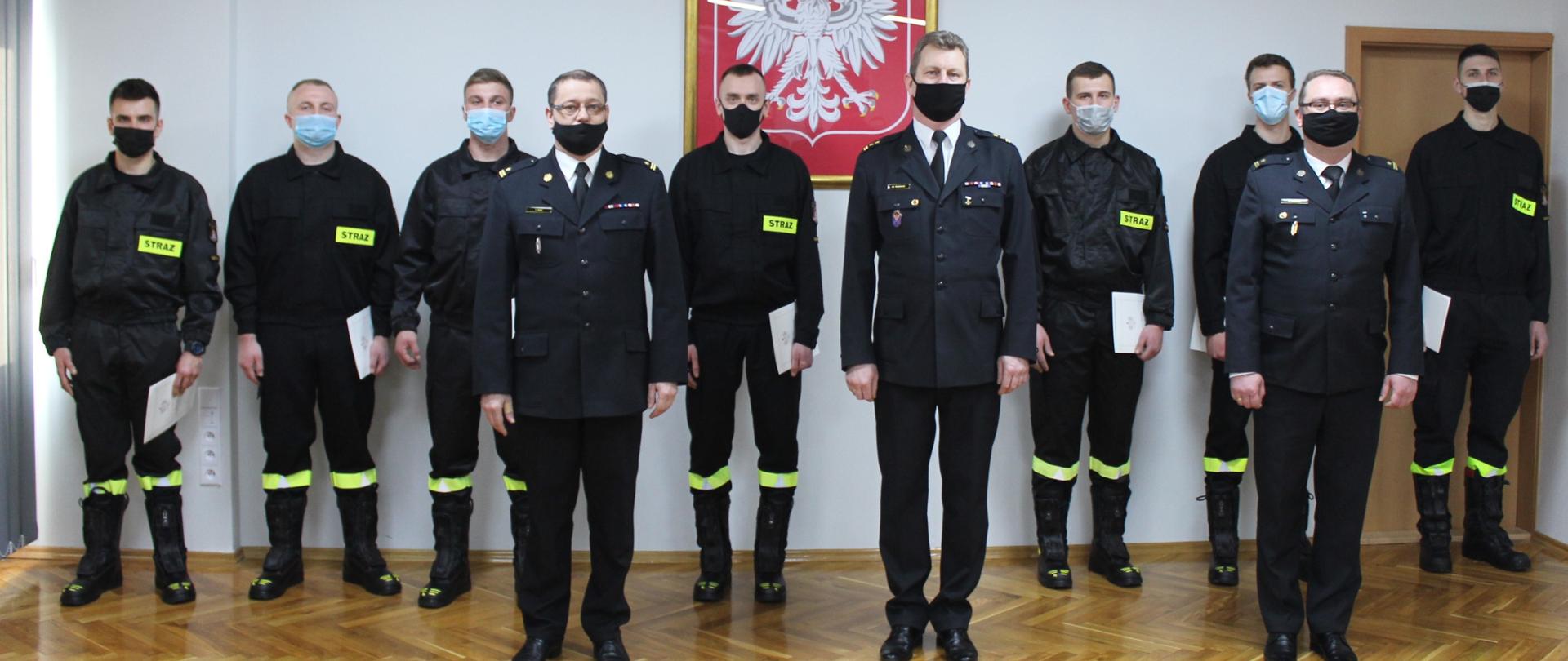 Zdjęcie przedstawia funkcjonariuszy Państwowej Straży Pożarnej podczas uroczystości ślubowania. Na zdjęciu znajduję się kierownictwo komendy w umundurowaniu wyjściowym oraz funkcjonariusze po ślubowaniu - 7 osób. Wszyscy funkcjonariusze są w maseczkach ochronnych. Zdjęcie wykonano na tle ściany na której wisi Godło Państwowe. 