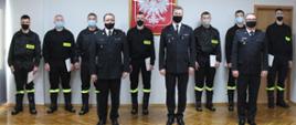 Zdjęcie przedstawia funkcjonariuszy Państwowej Straży Pożarnej podczas uroczystości ślubowania. Na zdjęciu znajduję się kierownictwo komendy w umundurowaniu wyjściowym oraz funkcjonariusze po ślubowaniu - 7 osób. Wszyscy funkcjonariusze są w maseczkach ochronnych. Zdjęcie wykonano na tle ściany na której wisi Godło Państwowe. 