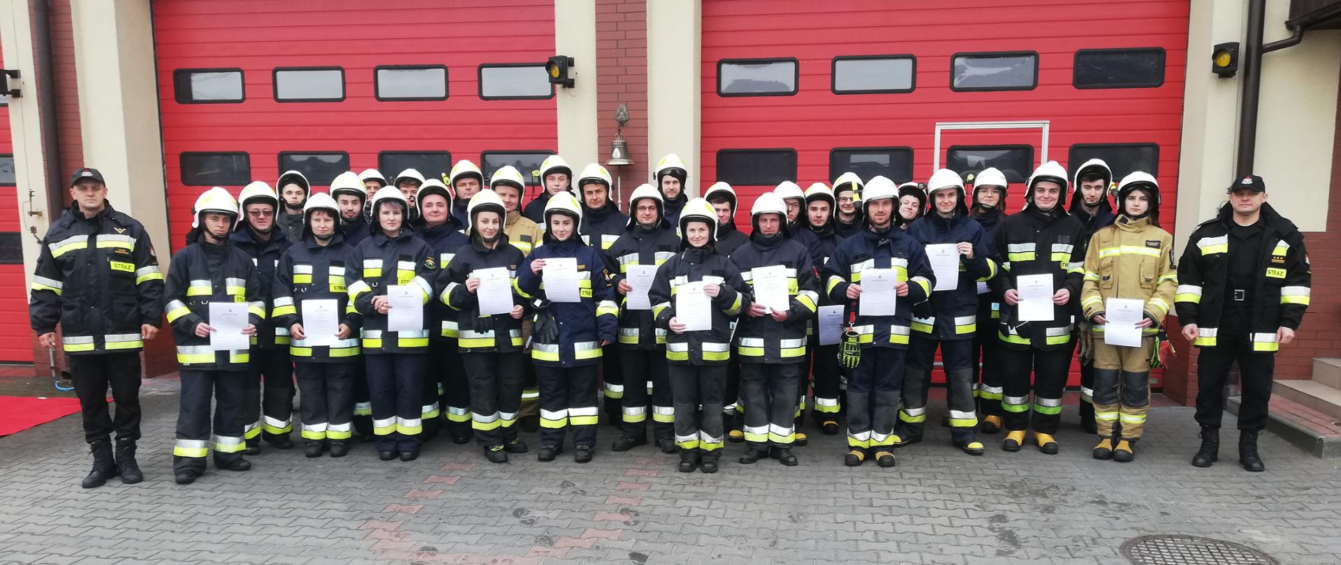 Kurs podstawowy OSP 2021 - zdjęcie grupowe po ukończeniu egzaminu. Strażacy stoją przed strażnicą Komendy trzymając w rękach dyplomy.