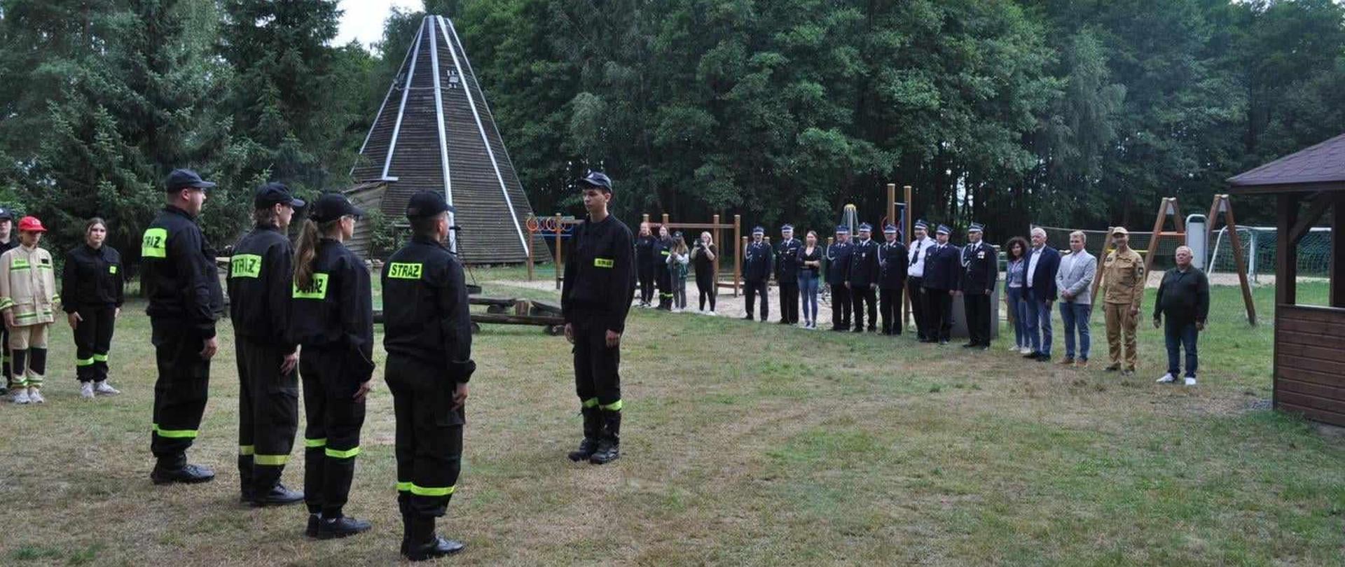 Zdjęcie przedstawia strażaków oraz dzieci i młodzież na wakacyjnym obozie strażackim.
W tle ośrodek wypoczynkowy.
