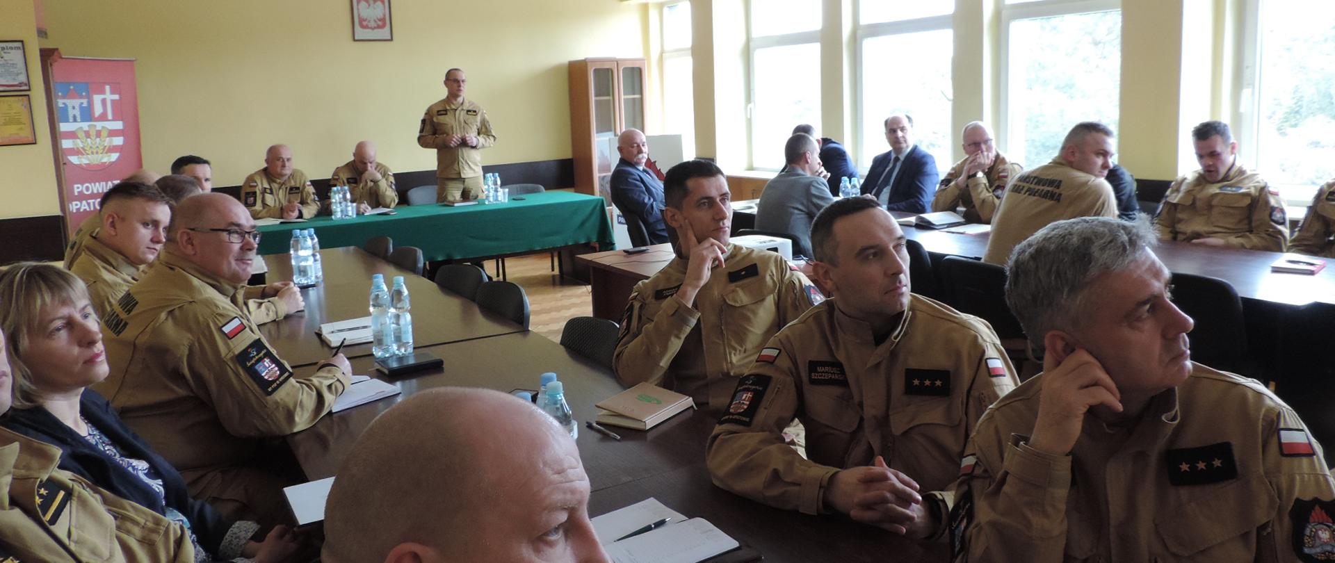 W tle Komendant KP PSP Opatów omawiający prezentację. Główny widok funkcjonariusze w umundurowaniu pisakowym oraz czarnym siedzący przy stole