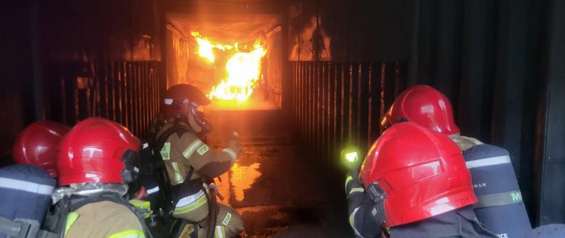 Zdjęcie przedstawia strażaków biorących udział w szkoleniu technik gaszenia pożarów wewnętrznych. W tle znajduje się kontener do ćwiczeń w którym w głębi pali się ogień a w nim znajdują się strażacy ubrani w nomexy, Chełmy i aparaty ochrony dróg oddechowych.