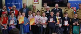 Na zdjęciu znajdują się dzieci z klas od 1 do 4 ze szkół podstawowych, które brały udział w turnieju. W rękach trzymają dyplomy i nagrody. Za dziećmi stoją przedstawiciele komisji egzaminacyjnej wraz ze starostą powiatowym i komendantem powiatowym Państwowej Straży Pożarnej w Dębicy.