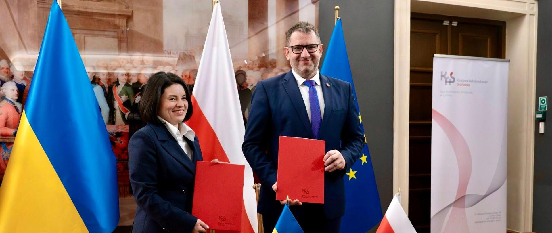 Na zdjęciu Szef KAS Marcin Łoboda oraz Lesya Karnaukh, p.o. Szefa Państwowej Służby Podatkowej Ukrainy, prezentują dopisane dokumenty, w tle flagi Polski i Ukrainy i UE