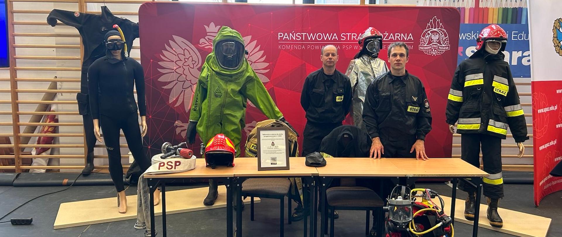 Udział strażaków Komendy Powiatowej PSP w Sokołowie Podlaskim w XIII Sokołowskich Targach Pracy, Edukacji i Kariery "Szarada" - na zdjęciu 2 strażaków z ekwipunkiem strażackim na sali gimnastycznej