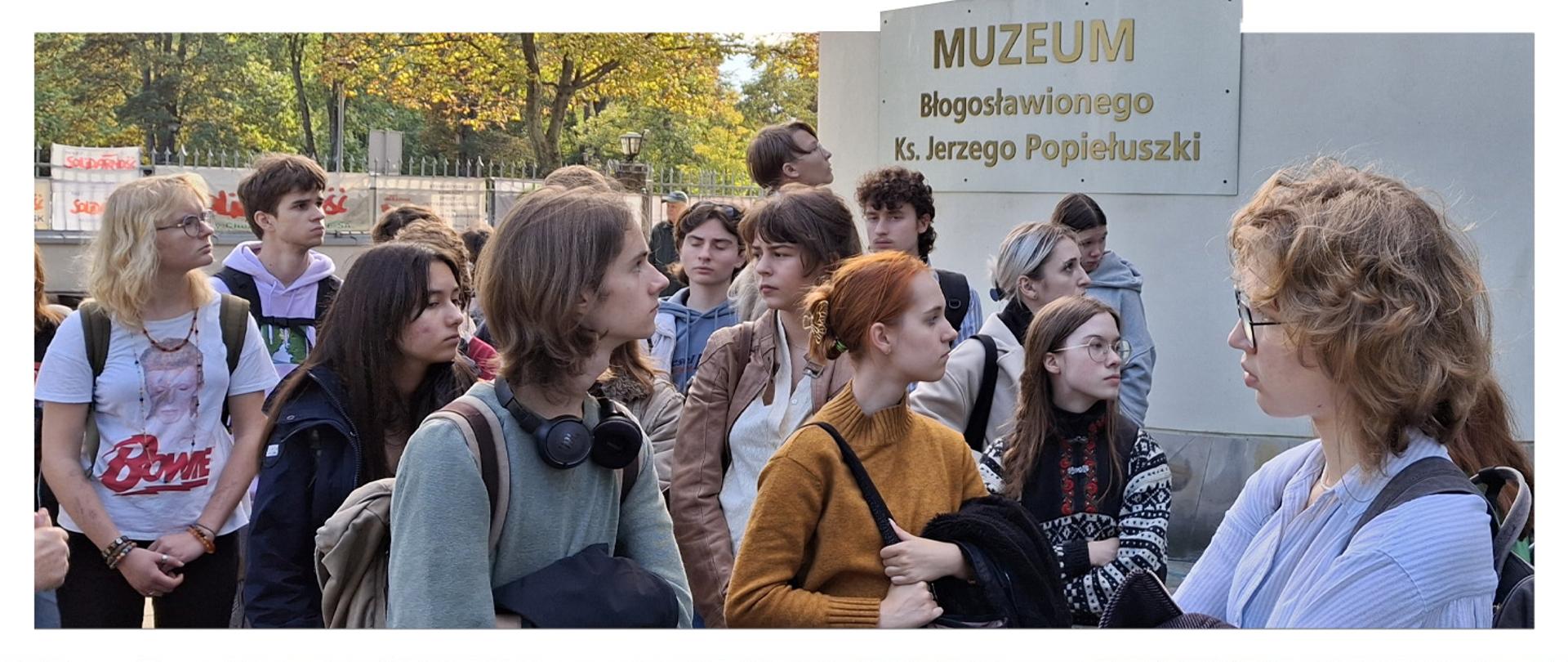 Uczniowie Liceum Plastycznego podczas wizyty w Muzeum Błogosławionego Księdza Jerzego Popiełuszki. Na pierwszym planie uczniowie, a w tle znajduje się tablica z nazwą muzeum.