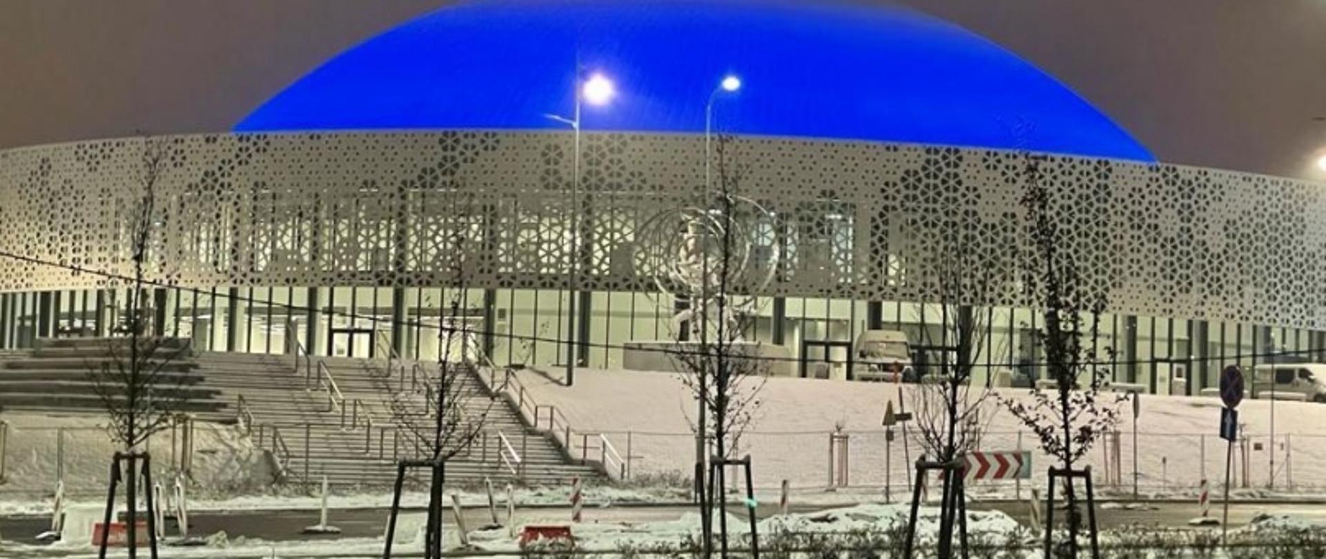 Nowoczesny budynek z kopułą i ażurową fasadą, oświetlony na niebiesko, w zimowej scenerii.