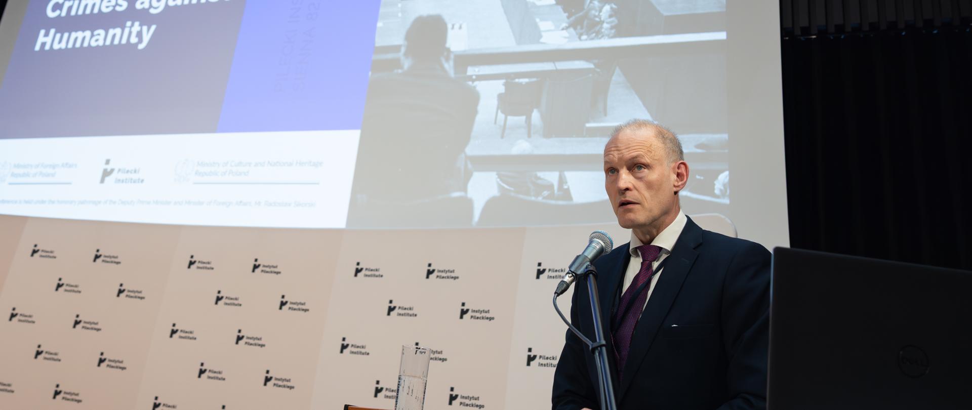 Deputy Minister of Foreign Affairs Wojciech Zajączkowski wearing a suit and a vine-colour tie speaks from the lectern on stage. There is a mike in front of him.