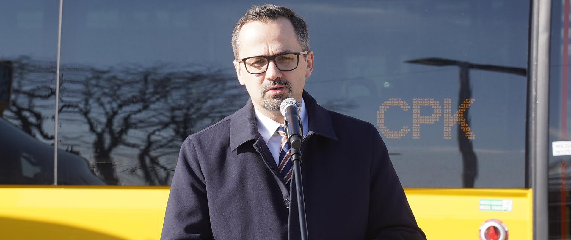 Wiceminister Marcin Horała stoi i mówi do mikrofonu. W tle za jego plecami stoi żółty autobus z ciemną, szklaną szybą.