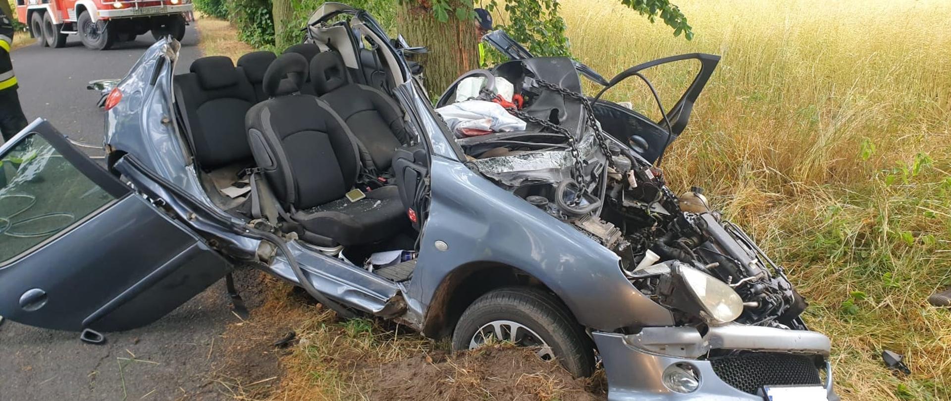 Zdjęcie na pierwszym planie przedstawia rozbity samochód marki peugeot 206 z odciętym dachem, w tle pojazd strażacki.