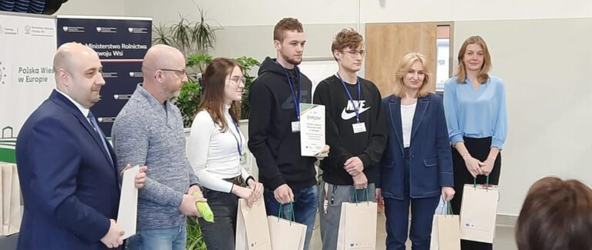 Grupa osób podczas ceremonii wręczenia nagród w pomieszczeniu. W centrum trójka młodych ludzi trzyma dyplom oraz torby upominkowe. Obok nich stoją przedstawiciele organizatorów w strojach wizytowych. W tle widoczna ścianka z logo Ministerstwa Rolnictwa i Rozwoju Wsi oraz napisem „Polska Wieś w Europie”.