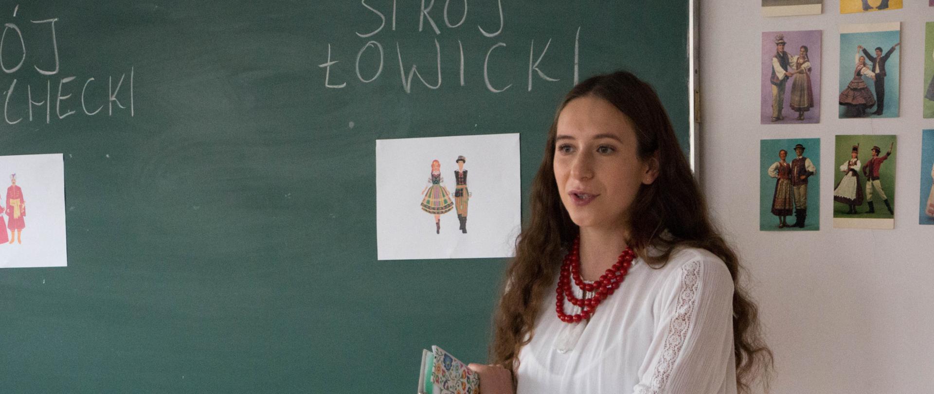 Słuchaczka Studium stoi i opowiada o swojej pracy dyplomowej pt. Polskie tańce narodowe. W tle obrazy przedstawiające stroje ludowe.