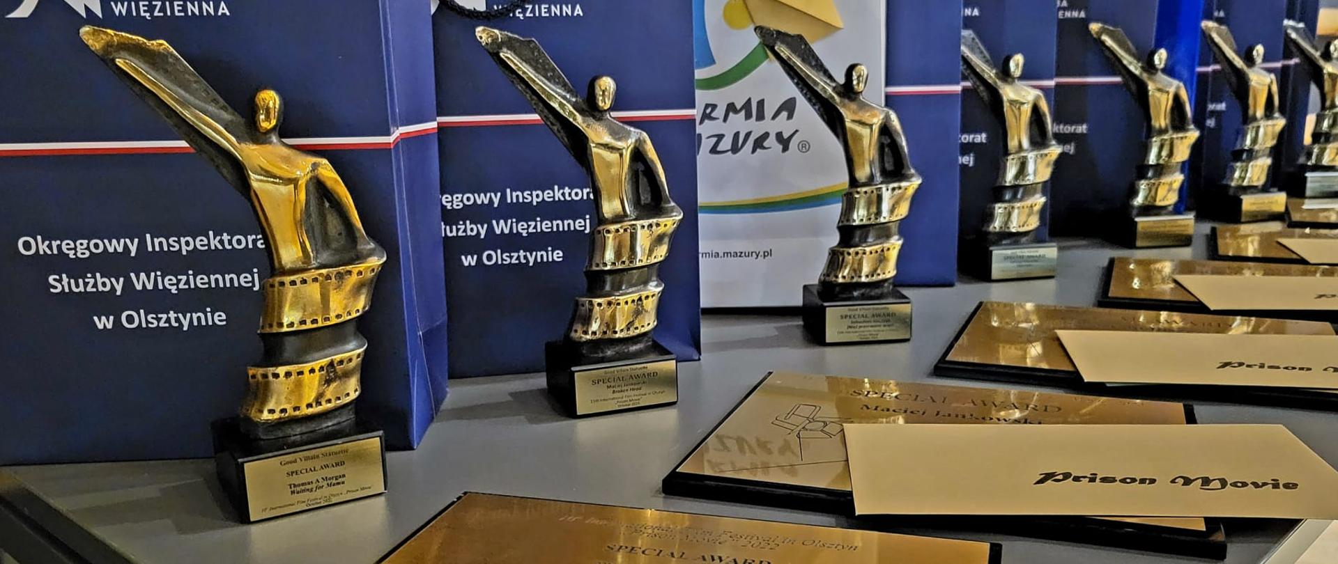 Na pierwszym planie widać koperty wraz z tabliczkami. Na drugim planie widnieją statuetki. 