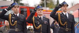 Trzej funkcjonariusze PSP w galowych mundurach oddają honory, salutując. W tle widoczne są wozy strażackie oraz strażackie sztandary.