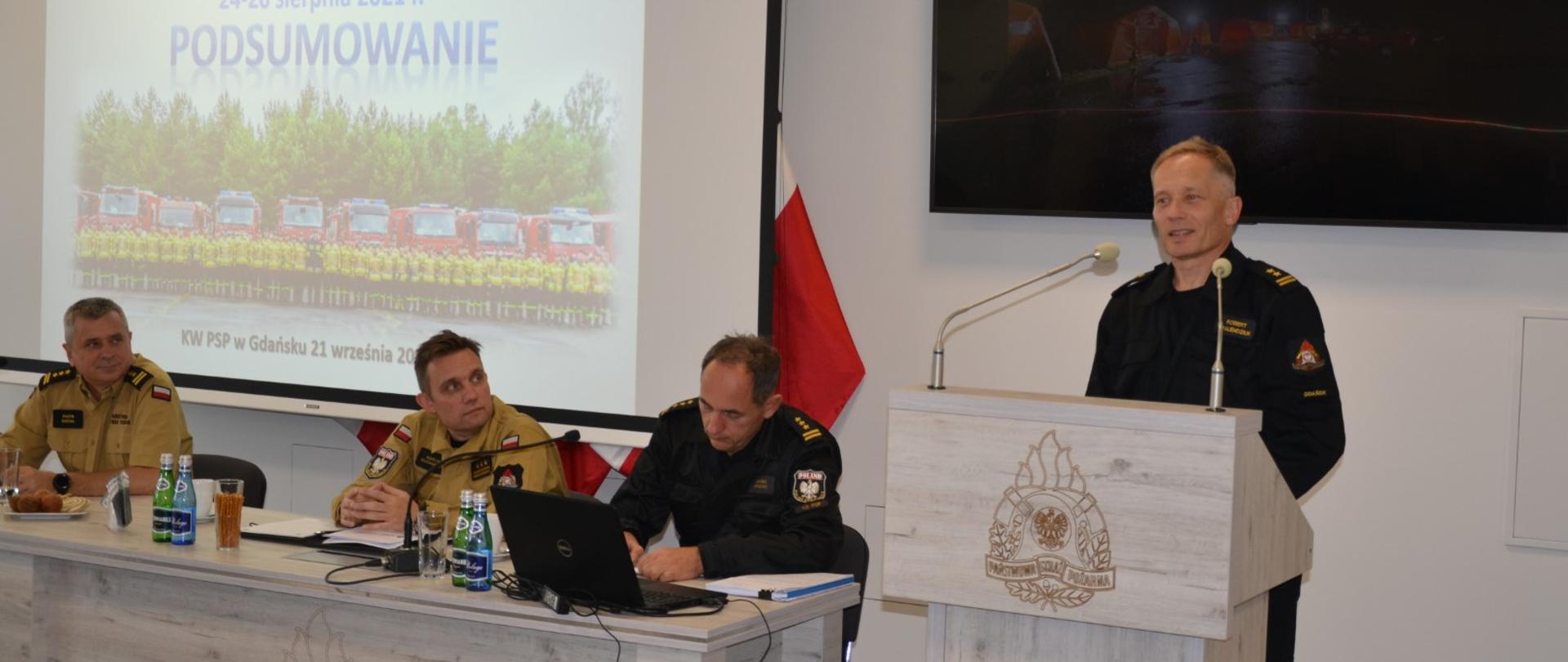 Strażak w obecności kadry kierowniczej Państwowej Straży Pożarnej omawia realizację ćwiczeń ratowniczych.