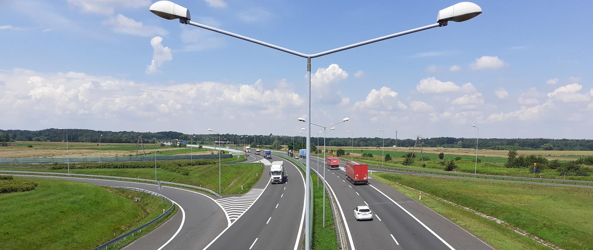Zdjęcie lotnicze autostrady A4. Na zdjęciu widoczne dwie jezdnie z poruszającymi się po niej pojazdami. Na środku zdjęcia widoczny pas rozdziału z zabudowanymi latarniami oświetlenia drogowego.
