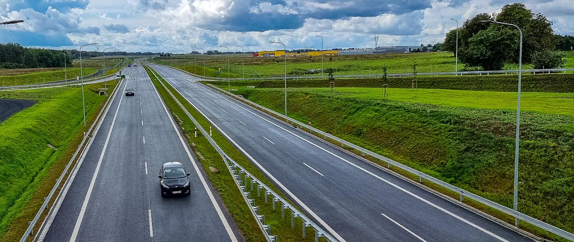 Dwujezdniowa trasa przedzielona pasem zieleni. Pochmurne niebo. Dookoła drogi dużo terenu zielonego.