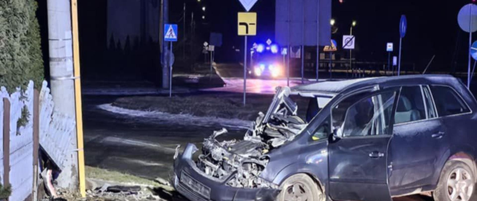 Nocna scena wypadku drogowego. Na pierwszym planie stoi mocno zniszczony samochód osobowy — przód pojazdu jest całkowicie rozbity, maska wyrwana i odsłonięta komora silnika. W tle stoi oświetlony pojazd strażacki z włączonymi światłami błyskowymi. Droga jest częściowo zablokowana, na jezdni widać rozsypane elementy pojazdu i ślady działań ratowniczych.