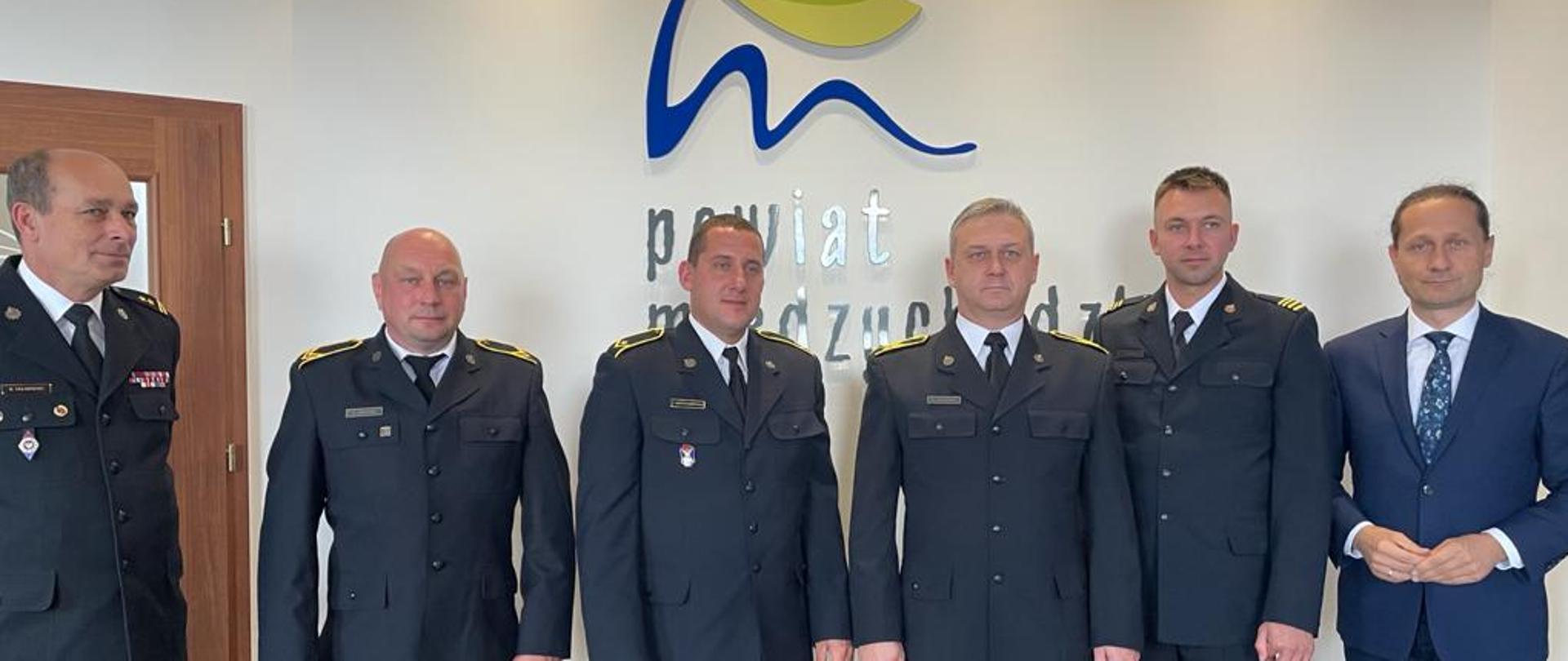 Na zdjęciu znajduje się 4 strażaków, którzy byli na misji w Grecji, obok Komendant Powiatowy PSP i Starosta Międzychodzki. W tle logo powiatu międzychodzkiego