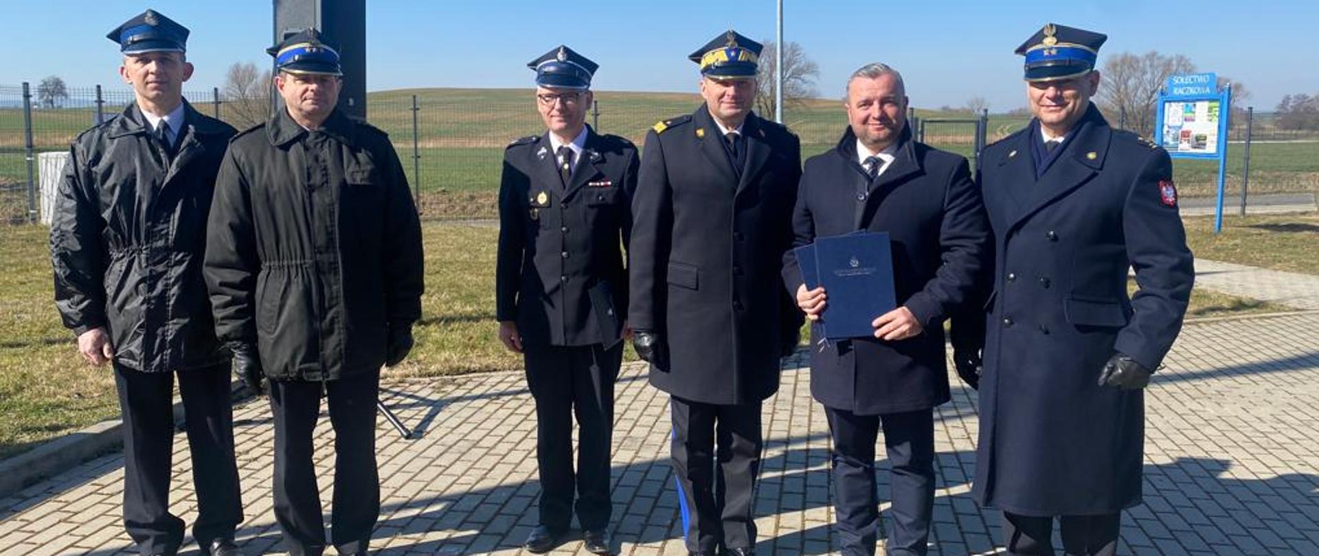 Obraz przedstawia przedstawicieli OSP Lipa i OSP Mysłów w towarzystwie Zastępcy Komendanta Głównego, Komendanta Wojewódzkiego, Komendanta Powiatowego i Burmistrza Bolkowa po wręczeniu promes na zakup samochodów pożarniczych. Pora dzienna.