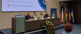 Przedstawiciele organizatorów programu - cztery osoby siedzą przy stole prezydialnym, nad nimi ekran multimedialny, obok stołu flagi państw uczestniczących w programie