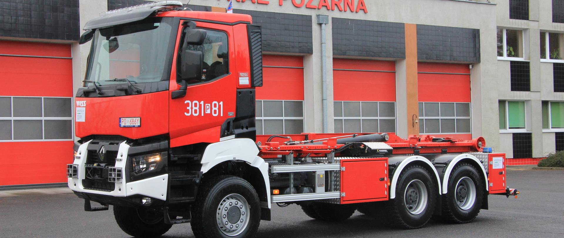 Samochód ciężarowy przeznaczony do przenoszenia kontenerów stoi na placu na tle budynku komendy. Widok z boku.